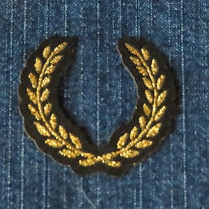 May include: A gold laurel wreath embroidered patch on a blue denim background.