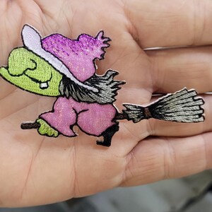 May include: An embroidered patch featuring a green-skinned character wearing a purple hat and pink outfit, riding a broomstick. The design includes black outlines, a light purple hat brim, and a grey broom with a brown handle, all with a glittery finish.