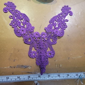Könnte beinhalten: Eine detaillierte, lila Spitzenapplikation mit einem Blumen- und Blätterdesign. Die Applikation ist symmetrisch, mit einem zentralen Blumenmotiv und sich erstreckenden, gebogenen Abschnitten. Die Spitze hat einen leuchtenden Lilaton, und das Design ist aufwendig und filigran.