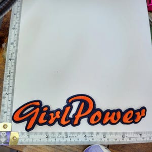 May include: Orange and black felt patch with the text "Girl Power" in a stylized font.