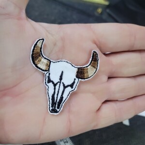 May include: Embroidered patch featuring a white bull skull with black outlining and brown and gold horns. The patch is designed with a detailed, stitched appearance, suitable for application on clothing or accessories.