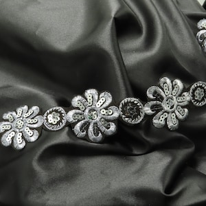 May include: Black satin fabric with a floral trim. The trim features embroidered flowers with silver sequins and beads. The fabric has a smooth, shiny texture and is draped in soft folds.