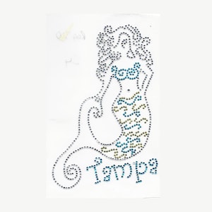 Peut inclure: Un design de sirène en strass avec une queue turquoise et or et le mot "Tampa" en dessous.