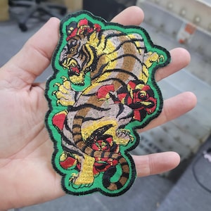 May include: Embroidered patch featuring a detailed tiger design with red roses. The tiger has brown and yellow stripes, a red mouth, and is set against a green border. The patch is likely for clothing or accessories.