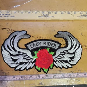 Op de afbeelding: Geborduurde patch met de tekst "LADY RIDER" boven een rode roos en groene bladeren, geflankeerd door gestileerde witte en grijze vleugels. De patch is zwart met een witte rand.