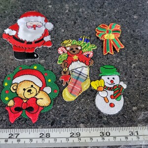 May include: A collection of embroidered Christmas patches featuring Santa Claus, a snowman, teddy bears, and a decorative bow. The patches are in various colors, including red, green, white, and gold. The patches are approximately 1 to 2 inches in size.