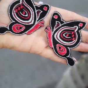 May include: Two embroidered butterfly patches featuring a black base with vibrant red and silver thread detailing. The butterfly designs have intricate patterns on the wings, including red accents and silver outlines, creating a distinct contrast.