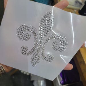 Pode incluir: Uma folha de plástico transparente com um design de flor-de-lis de strass prateados. O design é composto por inúmeros strass pequenos e redondos dispostos em forma de flor-de-lis estilizada. Os strass brilham contra o fundo branco.