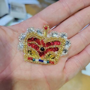 May include: A decorative crown patch, intricately detailed with gold thread, red sequins, and silver beads. The crown has a central black gem and a small red spire. The design is finished with a white lace trim, creating a regal appearance.