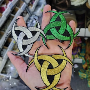 May include: Embroidered patches in silver, green, and gold, each featuring a stylized triquetra knot design with black accents. The patches are likely for clothing or accessories, offering a decorative element.