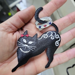 May include: Black cat patch with white embroidered details, a red bow, and a silver ring with red and blue stars. The cat has a smiling face and a curled tail. The patch is likely for clothing or accessories.
