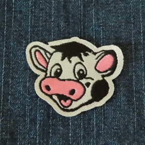 May include: Embroidered patch featuring a cartoon cow face with a white background, black spots, and pink details. The cow has a smiling expression with its tongue sticking out. The patch is outlined in white and is set against a blue denim background.