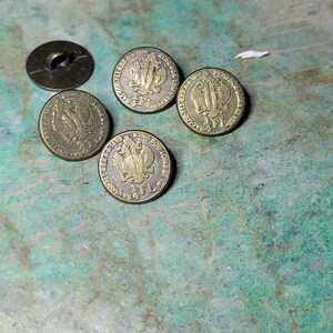 Puede incluir: Un conjunto de botones antiguos de latón y un botón de caña. Los botones redondos presentan un diseño en relieve con un escudo y el texto "4 FL". Los botones tienen un aspecto vintage y envejecido.