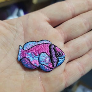 May include: Embroidered patch in the shape of a fish. The fish has a pink body with blue and purple accents. The patch is outlined in black.
