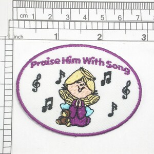 May include: A white oval embroidered patch with purple stitching. The patch features a cartoon girl with blonde hair and a purple dress singing with her hands clasped. The text "Praise Him With Song" is written in a purple font around the girl. Musical notes are embroidered around the girl.