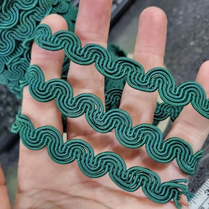 Pode incluir: Guarnição decorativa verde esmeralda com um design ondulado e em laço. A guarnição é feita de um material tecido. Ideal para costura e artesanato, adiciona um elemento decorativo aos projetos. Vista de perto.