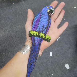 May include: Embroidered patch of a blue and purple macaw parrot perched on a green branch. The bird has a yellow eye and a black and white beak. The patch is detailed with various shades of blue, purple, and green.
