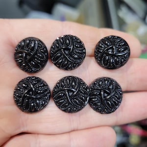 May include: Six black, round buttons with a detailed, textured design. Each button features a central, stylized floral or geometric pattern surrounded by a textured border. The buttons are made of a shiny, dark material.