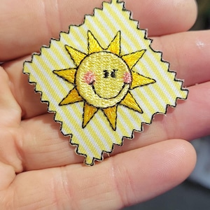 May include: A square embroidered patch with pinked edges featuring a yellow and white diagonal striped background. Centered on the patch is a yellow sun with a black embroidered smiling face, closed eyes, and small pink circular cheeks.