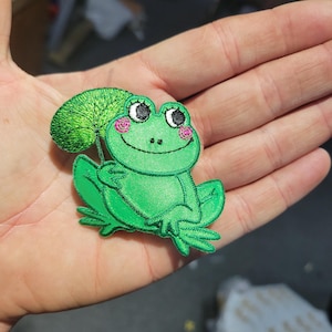 May include: A green embroidered frog patch with a smiling face, holding a green leaf. The frog has black and white eyes, pink cheeks, and is set against a neutral background.