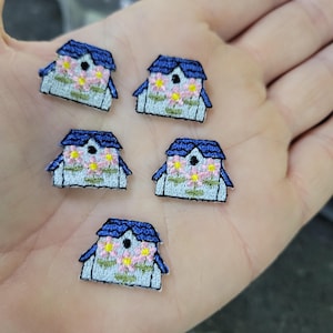 Könnte beinhalten: Fünf gestickte Aufnäher in Form kleiner Häuser. Jeder Aufnäher hat ein blaues Dach, weiße Wände und rosa und gelbe florale Details. Die Aufnäher sind klein und detailliert, mit einem genähten Aussehen.