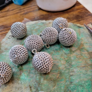 May include: Close-up of eight silver metal balls with a woven design. Each ball has a small loop attached.