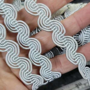 May include: Close-up of a light silver wavy trim. The trim has a looped design, creating a continuous wave pattern. The trim is held in a hand, showing the texture and detail of the material. The trim is likely used for sewing or crafting projects.