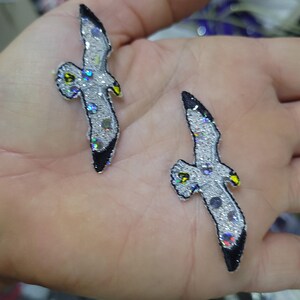 May include: Two decorative seagull patches. The patches are silver with iridescent sequins and black accents. The seagulls have yellow beaks and are in a flying position. These patches could be used to embellish clothing or accessories.