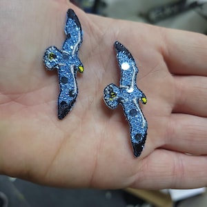 May include: Two blue glitter bird patches with black and yellow accents. The patches are in the shape of a bird in flight. The patches are held in a person's hand.