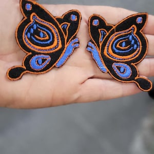 May include: Two embroidered butterfly patches featuring a black base with vibrant orange and blue thread detailing. The butterfly designs showcase intricate patterns with blue accents on the wings and orange outlines, creating a bold contrast.