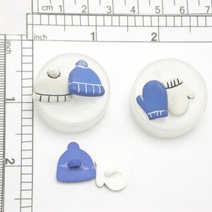May include: Miniature winter-themed ornaments featuring a blue and white hat, a blue and white mitten, and a blue hat and white mitten. The ornaments are on a white surface, with a ruler showing measurements in cm and inches.