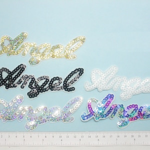 Peut inclure: Cinq patchs thermocollants avec le mot "Angel" écrit en paillettes. Les patchs sont de différentes couleurs : or, noir, blanc, iridescent et arc-en-ciel.