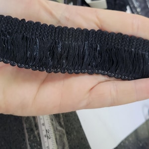 May include: Close-up of a black fringe trim. The trim features a woven top edge with a series of hanging fringe. The fringe is a solid black color. The trim is held in a hand.