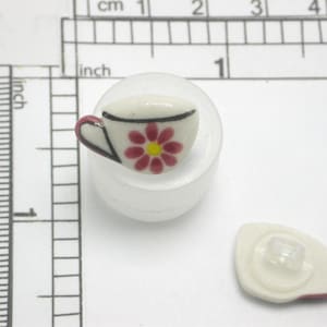 Könnte beinhalten: Ein kleiner Keramikknopf in Form einer Teetasse mit weißem Sockel, einem rot-gelben Blumenmuster und einem schwarzen Henkel. Der Knopf hat einen Durchmesser von ca. 2 cm. Ein passendes Knopfrückteil ist ebenfalls sichtbar.