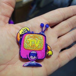 May include: A colorful embroidered patch in the shape of a retro television set. The TV is pink with a green screen displaying the word "TV" in yellow. It has blue antennae and yellow arms and hands.
