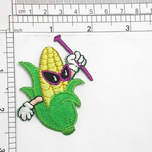 May include: An embroidered patch of a cartoon corn cob wearing sunglasses and holding a purple stick. The corn cob is yellow with green leaves and white gloves.