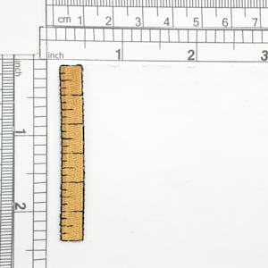 Op de afbeelding: Een kleine, bruine stoffen liniaal met zwarte markeringen. De liniaal is ongeveer 5 cm lang.