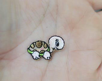 May include: Embroidered turtle patch with a white head, tan shell, and green accents. The turtle has a friendly expression and is likely for clothing or craft projects. The patch is small and detailed.