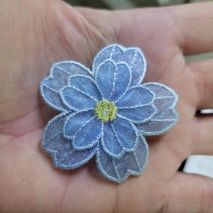 May include: A delicate, light blue embroidered flower with multiple petals and a yellow center. The flower is made of a sheer fabric and has a detailed, textured appearance. The floral design is likely for embellishment or crafting.