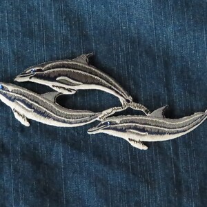 Peut inclure: Écusson brodé représentant trois dauphins gris et blancs, avec des yeux bleus. Les dauphins sont disposés dans un motif fluide, sur un fond bleu texturé, adapté aux vêtements ou à l'artisanat.