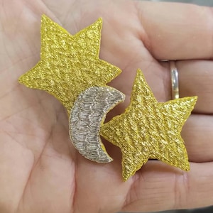 May include: Two embroidered star patches in a bright metallic gold thread, featuring a textured finish. A single silver crescent moon patch with a matching textured thread pattern is positioned between the two gold stars, creating a celestial set.