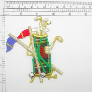 Könnte beinhalten: Eine grüne, rote und weiße karierte Golftasche mit goldenen Verzierungen und einer blau-roten Flagge. Die Tasche ist mit Golfschlägern gefüllt und hat einen goldenen Boden.