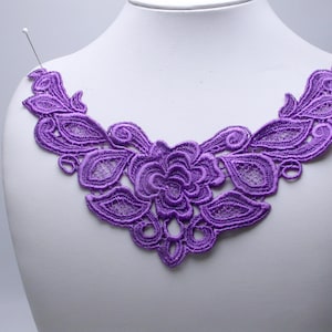 Puede incluir: Una aplicación de cuello de encaje en color morado vibrante con un intrincado patrón de bordado de flores y hojas. El diseño es simétrico y presenta un motivo central de rosa flanqueado por follaje y detalles calados con alfileres.