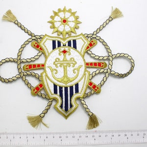 May include: A gold embroidered patch with a white and blue striped shield, an anchor, and two crossed swords. The shield is surrounded by a gold rope with tassels. The patch is approximately 10 inches wide.