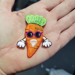 May include: Embroidered orange carrot patch with green leafy top, purple sunglasses, and white gloves. The carrot has a smiling face and is a fun, whimsical design. This patch is perfect for adding to clothing or accessories.