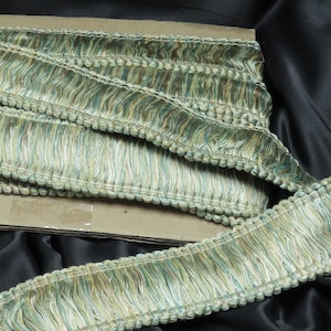 May include: A close-up of a green and brown fringe trim with a beaded edge. The trim is folded in half and is lying on a black fabric surface.