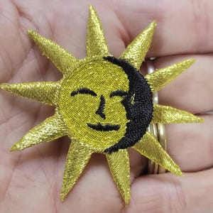 May include: A sun and moon embroidered patch featuring a metallic gold thread sun with sharp rays and a black thread crescent moon overlay. The sun has a simple stitched face with closed eyes and a small mouth, creating a celestial design.