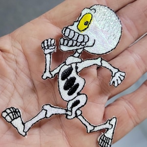 May include: An embroidered patch featuring a white skeleton in a running pose with black outlines and details. The skeleton has a large, round head with a single prominent yellow eye and a wide, toothy grin. The design is rendered in white thread.