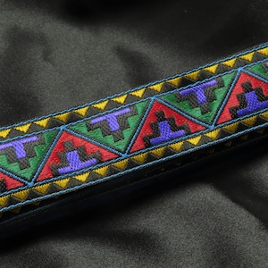 May include: A decorative trim with a geometric pattern. The trim features a repeating design of triangles and stepped shapes in red, green, purple, and black, outlined with yellow and blue stitching. The trim is on a dark background.