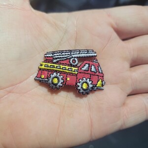 May include: Embroidered patch of a red fire truck with a silver ladder on top. The fire truck has yellow and black detailing on the wheels and body. The patch is likely for clothing or accessories.